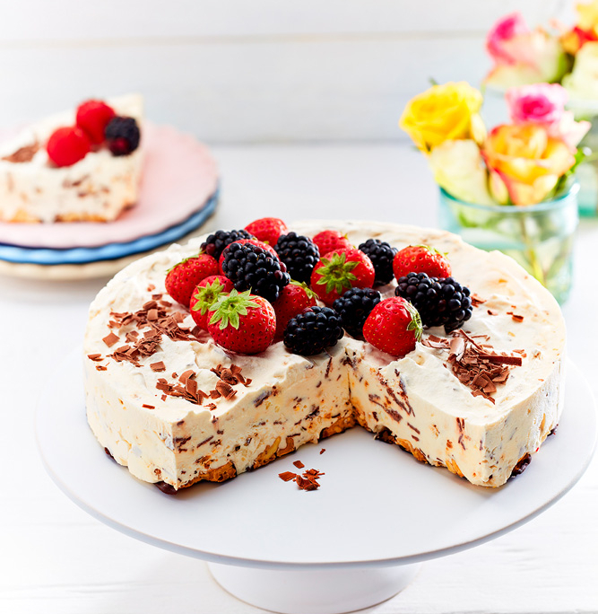 Eine dekadente Eistorte, gefertigt von einer talentierten Foodstylistin, ist mit frischen Erdbeeren und Brombeeren belegt und steht auf einem weißen Tortenständer. Ein Stück wurde entfernt, so dass das attraktive Innere mit Schokoladenraspeln zum Vorschein kommt. Im Hintergrund sind eine Vase mit bunten Blumen und ein Teller mit einem Stück Torte zu sehen – ein wahres Meisterwerk der Foodfotografie.