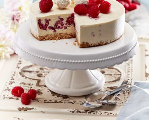 Einw weiße Eistorte mit einer Himbeer im Inneren steht auf einem weißen Tortenständer und verkörpert exquisite Food-Fotografie. Der Kuchen ist mit ganzen Himbeeren und weißen Schokoladentrüffeln garniert. Es sind Stücke ausgeschnitten und eine antike, spitzenartige Unterlage, Löffel und eine Serviette schmücken den rustikalen Holztisch.