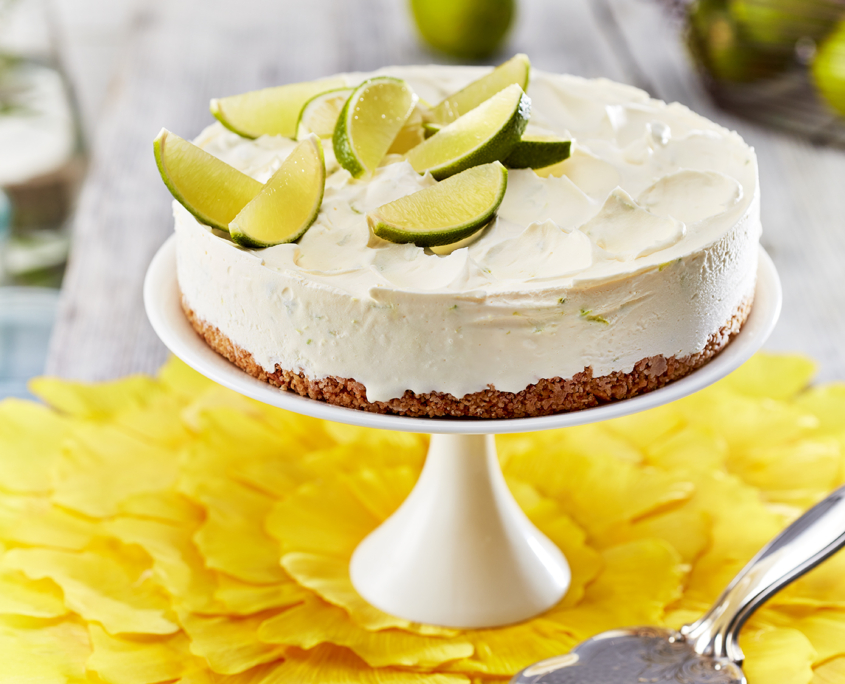 Ein Limetten-Eistorte mit frischen Limettenspalten steht auf einem weißen Tortenständer auf einem mit gelben Blütenblättern geschmückten Tisch. Die Szene wurde von talentierten Food-Fotografen aufgenommen und umfasst einen silbernen Servierspatel und weitere Limetten, mit einem verschwommenen Hintergrund, auf dem weitere Limetten und gelbe Blumen in einer Vase zu sehen sind.
