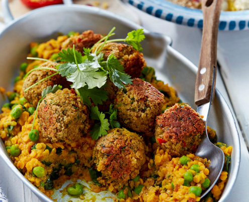 prizibylla-fotograf-levante-bunte-falaffel-auf-spinatlinsen Eine Servierplatte mit Falafelbällchen, garniert mit Koriander, auf einem Bett aus Roten Linsen, Spinat und grünen Erbsen. Ein Löffel liegt am Rand der Platte. Im Hintergrund sind Teller mit bunten Salaten und verschiedenen Toppings zu sehen – eine perfekte Aufnahme für jeden Food-Fotografie-Enthusiasten.