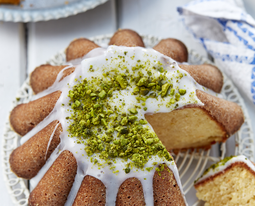 prizibylla-fotograf-levante-getraenkter-zitronenkuchen Ein mit weißer Glasur überzogener und mit gehackten Pistazien bestreuter Zitronenkuchen liegt auf einem weißen Tortengitter. Ein Stück des Kuchens wurde abgeschnitten, so dass das helle, saftige Innere sichtbar wird. Im Hintergrund sind ein weiterer Kuchen und eine blau-weiße Serviette zu sehen, die die Essenz exquisiter Foodfotografie einfangen.