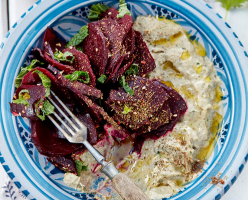 przibylla-fotograf-levante-baba-ganoush-mit-ofen-bete Auf einem blau-weißen Teller ist eine Portion Baba Ganoush mit Roter Bete, garniert mit Kräutern und Dukah, beträufelt mit Olivenöl. Eine exquisite Food-Fotografie fängt eine Gabel auf dem Teller ein, und darunter ist teilweise ein gemustertes Tuch zu sehen.