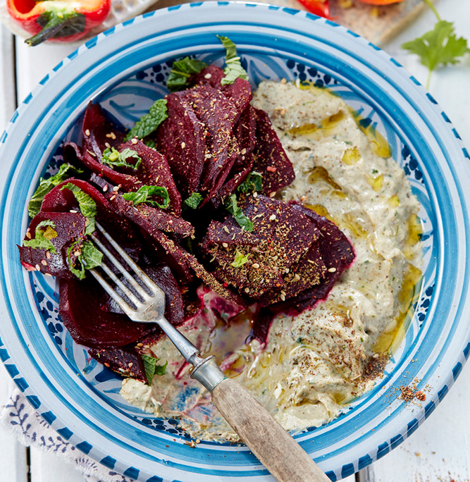 przibylla-fotograf-levante-baba-ganoush-mit-ofen-bete Auf einem blau-weißen Teller ist eine Portion Baba Ganoush mit Roter Bete, garniert mit Kräutern und Dukah, beträufelt mit Olivenöl. Eine exquisite Food-Fotografie fängt eine Gabel auf dem Teller ein, und darunter ist teilweise ein gemustertes Tuch zu sehen.
