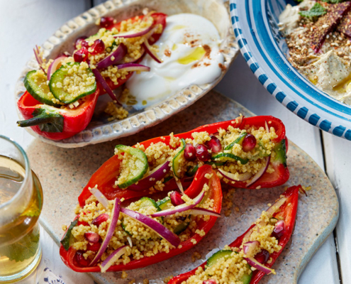 przibylla-fotograf-levante-couscous-salat-mit-gebackener-paprika Eine wunderschön gedeckte Tischplatte zeigt die Kunstfertigkeit eines Foodstylisten. Darin sind rote Paprikaschiffchen zu sehen, die mit Couscous, Gurke, roten Zwiebeln und Granatapfelkernen gefüllt sind. Daneben steht ein Schälchen mit cremigem weißen Dip und einer gefüllten Spitzpaprika, eine Schale mit Falafel und buntem Reis im Hintergrund und ein blauer Teller mit Baba Ganouch und Roter Bete sowie ein Glas Weißwein. Foodfotografie vom Feinsten!