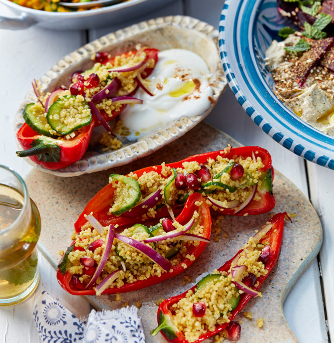 przibylla-fotograf-levante-couscous-salat-mit-gebackener-paprika Eine wunderschön gedeckte Tischplatte zeigt die Kunstfertigkeit eines Foodstylisten. Darin sind rote Paprikaschiffchen zu sehen, die mit Couscous, Gurke, roten Zwiebeln und Granatapfelkernen gefüllt sind. Daneben steht ein Schälchen mit cremigem weißen Dip und einer gefüllten Spitzpaprika, eine Schale mit Falafel und buntem Reis im Hintergrund und ein blauer Teller mit Baba Ganouch und Roter Bete sowie ein Glas Weißwein. Foodfotografie vom Feinsten!