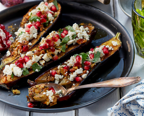 przibylla-fotograf-levante-gebackene-auberginen-mit-feta Ein schwarzer Teller mit halbierten gebackenen Auberginen, garniert mit zerbröseltem Feta, Granatapfelkernen und frischen Kräutern. Um den Teller herum stehen bunte Trinkgläser und weitere Teller mit verschiedenen Gerichten, alles auf einem weißen Holztisch gedeckt – die perfekte Bildsprache für jeden Food-Fotografen.