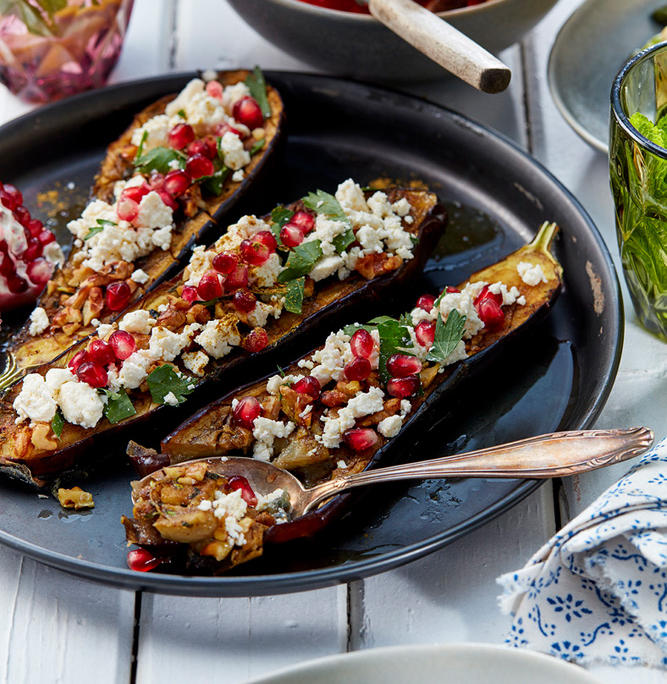 przibylla-fotograf-levante-gebackene-auberginen-mit-feta Ein schwarzer Teller mit halbierten gebackenen Auberginen, garniert mit zerbröseltem Feta, Granatapfelkernen und frischen Kräutern. Um den Teller herum stehen bunte Trinkgläser und weitere Teller mit verschiedenen Gerichten, alles auf einem weißen Holztisch gedeckt – die perfekte Bildsprache für jeden Food-Fotografen.