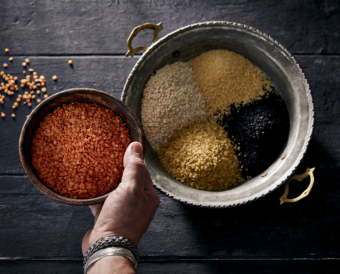 przibylla-fotograf-levante-huelsenfruechte-mood Eine Hand hält eine Schüssel mit roten Linsen über einem silbernen Tablett mit Griffen. Das Tablett, perfekt für Foodfotografie gestaltet, enthält verschiedene getrocknete Hülsenfrüchte, darunter schwarzen Quinoa, gelben Bulgur und Couscous, alles auf einer dunklen Holzoberfläche.