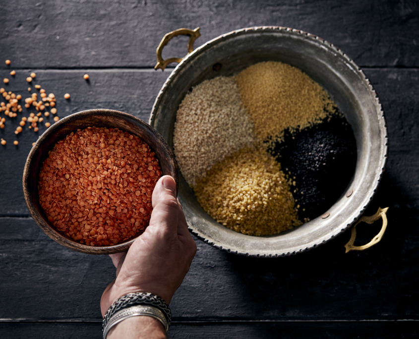 przibylla-fotograf-levante-huelsenfruechte-mood Eine Hand hält eine Schüssel mit roten Linsen über einem silbernen Tablett mit Griffen. Das Tablett, perfekt für Foodfotografie gestaltet, enthält verschiedene getrocknete Hülsenfrüchte, darunter schwarzen Quinoa, gelben Bulgur und Couscous, alles auf einer dunklen Holzoberfläche.