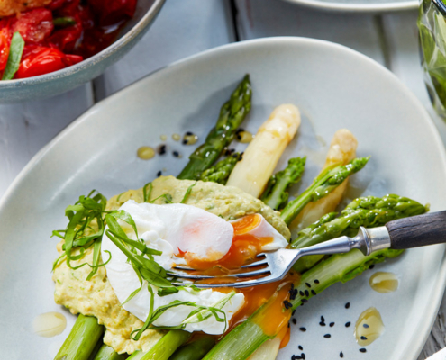 przibylla-fotograf-levante-hummus-mit-spargel-und-ei Ein Teller mit grünem und weißem Spargel, gekrönt von einem pochierten Ei mit flüssigem Eigelb, garniert mit einem gelb-grünen Dip. Der von einem erfahrenen Foodstylisten arrangierte Teller ist mit Microgreens garniert, wobei andere Gerichte und eine Serviette teilweise im Hintergrund sichtbar sind.