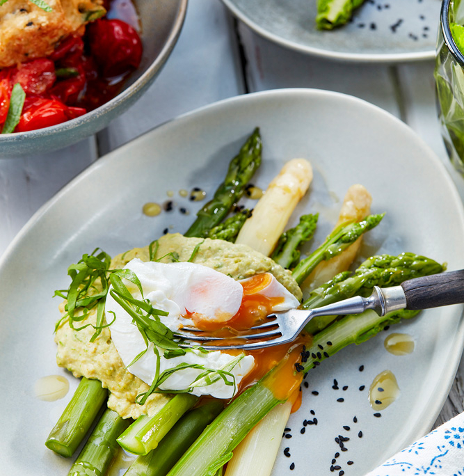 przibylla-fotograf-levante-hummus-mit-spargel-und-ei Ein Teller mit grünem und weißem Spargel, gekrönt von einem pochierten Ei mit flüssigem Eigelb, garniert mit einem gelb-grünen Dip. Der von einem erfahrenen Foodstylisten arrangierte Teller ist mit Microgreens garniert, wobei andere Gerichte und eine Serviette teilweise im Hintergrund sichtbar sind.