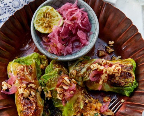 przibylla-fotograf-levante-kartoffel-blumenkohl-roulade-mit-zwiebelconfit Auf einem bronzenen Teller liegen gefüllte Kohlrouladen, garniert mit gehackten Nüssen und rotem Zwiebelnkonfit, perfekt für Food-Fotografen, die die filigranen Details einfangen möchten. Eine halbierte Limette und eine kleine Schale mit eingelegten Zwiebeln stehen daneben. Im Hintergrund sieht man einen weißen Teller mit geschnittenem Fladenbrot, das mit Labneh, Gurken und Rucola belegt und ein Glas Limettenwasser.