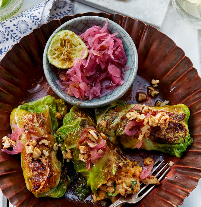 przibylla-fotograf-levante-kartoffel-blumenkohl-roulade-mit-zwiebelconfit Auf einem bronzenen Teller liegen gefüllte Kohlrouladen, garniert mit gehackten Nüssen und rotem Zwiebelnkonfit, perfekt für Food-Fotografen, die die filigranen Details einfangen möchten. Eine halbierte Limette und eine kleine Schale mit eingelegten Zwiebeln stehen daneben. Im Hintergrund sieht man einen weißen Teller mit geschnittenem Fladenbrot, das mit Labneh, Gurken und Rucola belegt und ein Glas Limettenwasser.