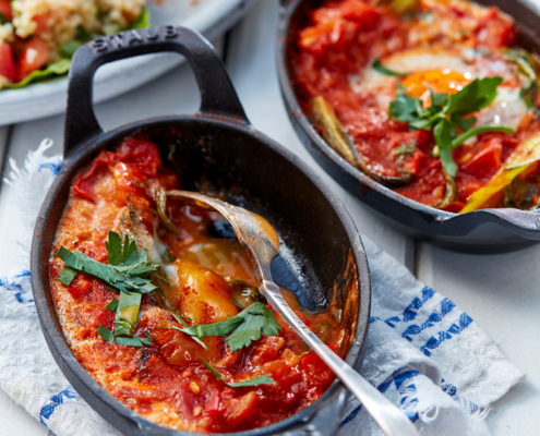 przibylla-fotograf-levante-mangold-shashuka Zwei kleine gusseiserne Pfännchen mit Shakshuka, einem nordafrikanischen Gericht aus Eiern in einer würzigen Tomatensauce, garniert mit frischer Petersilie, stehen auf einem weißen Tisch mit einem blau-weißen Tuch. In einer der Pfannen liegt ein Löffel. Im Hintergrund ein Teller mit Taboulé – aufgenommen von einem erfahrenen Food-Fotografen.