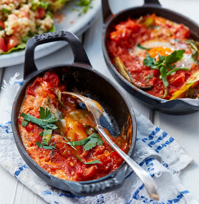 przibylla-fotograf-levante-mangold-shashuka Zwei kleine gusseiserne Pfännchen mit Shakshuka, einem nordafrikanischen Gericht aus Eiern in einer würzigen Tomatensauce, garniert mit frischer Petersilie, stehen auf einem weißen Tisch mit einem blau-weißen Tuch. In einer der Pfannen liegt ein Löffel. Im Hintergrund ein Teller mit Taboulé – aufgenommen von einem erfahrenen Food-Fotografen.