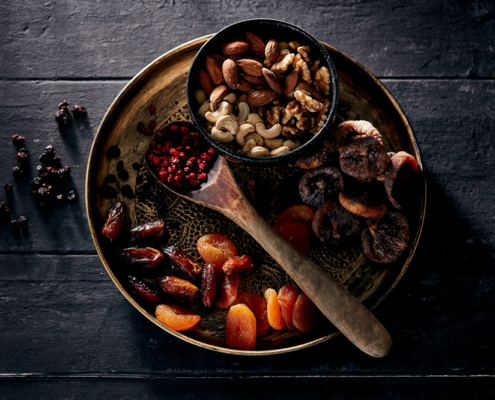 przibylla-fotograf-levante-nuesse-trockenfruechte-stilllife Eine Topshot auf einen runde Metallplatte, kunstvoll im Foodfotografie-Stil arrangiert, präsentiert eine Auswahl an Trockenfrüchten und Nüssen, darunter Datteln, Aprikosen, Feigen, Mandeln, Cashews und Walnüsse. Auf dem Teller liegt auch ein Holzlöffel mit roten Beeren. Verstreute Trockenbeeren zieren die dunkle Holzoberfläche.