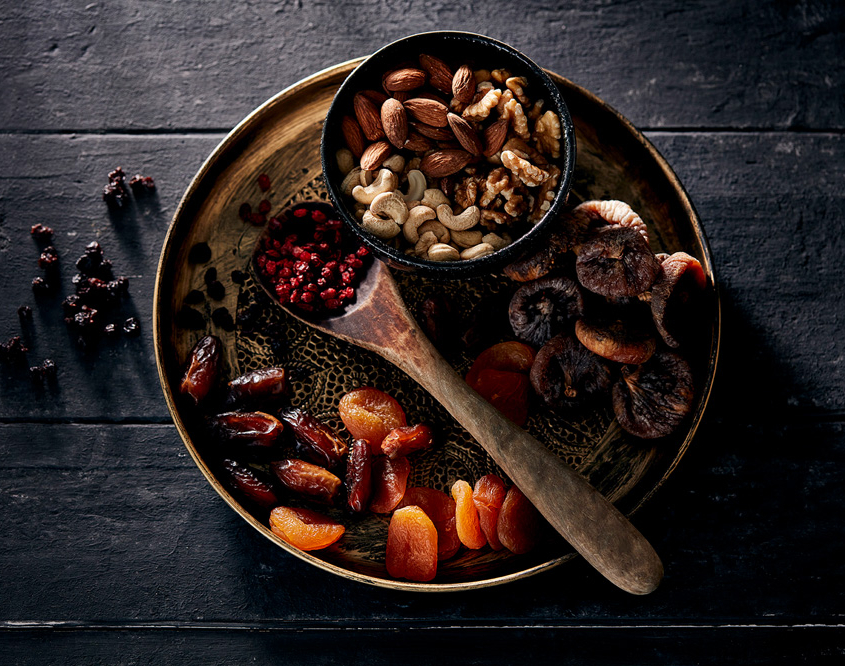 przibylla-fotograf-levante-nuesse-trockenfruechte-stilllife Eine Topshot auf einen runde Metallplatte, kunstvoll im Foodfotografie-Stil arrangiert, präsentiert eine Auswahl an Trockenfrüchten und Nüssen, darunter Datteln, Aprikosen, Feigen, Mandeln, Cashews und Walnüsse. Auf dem Teller liegt auch ein Holzlöffel mit roten Beeren. Verstreute Trockenbeeren zieren die dunkle Holzoberfläche.