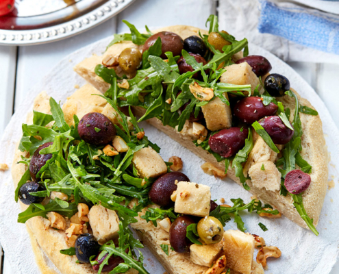 przibylla-fotograf-levante-oliven-brotsalat-mit-nuessen Ein rundes, flaches Focaccia-Brot, belegt mit Rucola, Oliven, gebackenen Brotwürfeln und Nüssen, ist in drei Stücke geschnitten. Im Hintergrund sind teilweise Teller mit geröstetem Gemüse, darunter Kartoffeln und Kirschtomaten, zu sehen – ein Beispiel für die exquisite Expertise eines Foodstylisten.