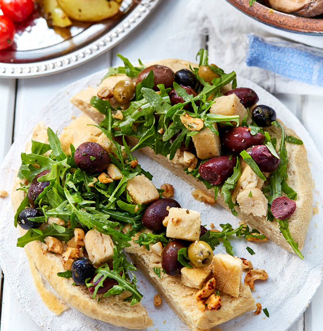 przibylla-fotograf-levante-oliven-brotsalat-mit-nuessen Ein rundes, flaches Focaccia-Brot, belegt mit Rucola, Oliven, gebackenen Brotwürfeln und Nüssen, ist in drei Stücke geschnitten. Im Hintergrund sind teilweise Teller mit geröstetem Gemüse, darunter Kartoffeln und Kirschtomaten, zu sehen – ein Beispiel für die exquisite Expertise eines Foodstylisten.