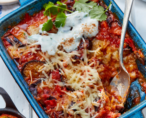 przibylla-fotograf-levante-orientalische-parmigiana-mit-kraeuterjoghurt Auf einer blauen Keramikschale liegt eine gebackener Parmigiana mit geschmolzenem Käse, Aubergine, Tomaten Soße und frischen Kräutern, festgehalten in atemberaubender Bildsprache. In der Schale liegt ein Metalllöffel. Daneben stehen eine Schüssel mit Joghurtsoße und ein Teller Taboulé auf einem weißen Holztisch.