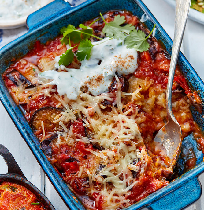 przibylla-fotograf-levante-orientalische-parmigiana-mit-kraeuterjoghurt Auf einer blauen Keramikschale liegt eine gebackener Parmigiana mit geschmolzenem Käse, Aubergine, Tomaten Soße und frischen Kräutern, festgehalten in atemberaubender Bildsprache. In der Schale liegt ein Metalllöffel. Daneben stehen eine Schüssel mit Joghurtsoße und ein Teller Taboulé auf einem weißen Holztisch.