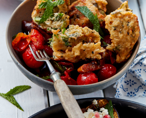 przibylla-fotograf-levante-reiskrapfen-mit-tomate-und-feigen In einer Schüssel liegen gebratene Reiskrapfen, garniert mit Minzeblättern, auf gerösteten Kirschtomaten und Paprika. In der Schüssel liegt eine Gabel. Im Hintergrund ist die Handschrift eines Foodstylisten in einem weiteren Gericht mit weißen und grünen Spargelstangen und pochierten Eiern zu erkennen. Im Vordergrund gefüllte Auberginen, garniert mit Feta und Granatapfel.