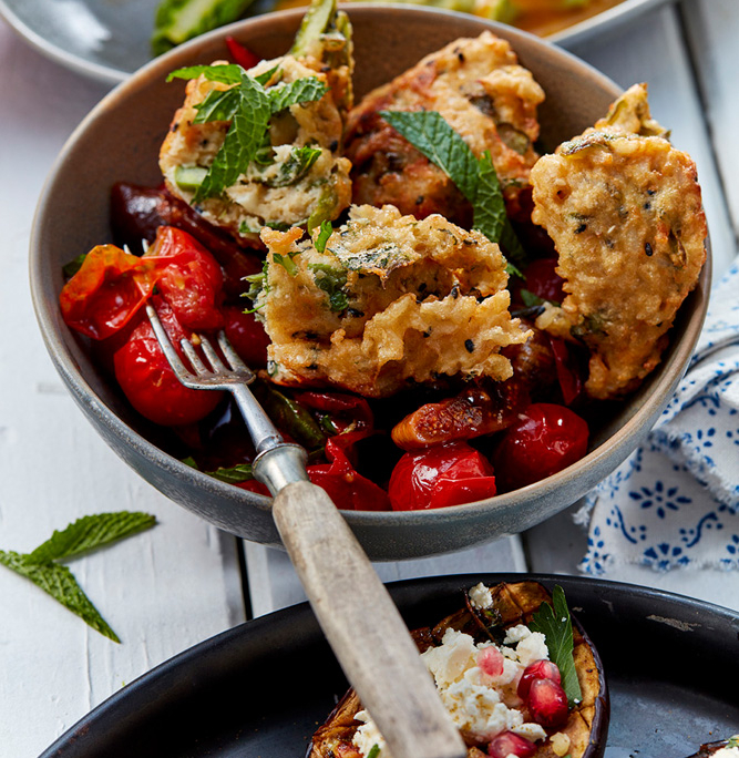 przibylla-fotograf-levante-reiskrapfen-mit-tomate-und-feigen In einer Schüssel liegen gebratene Reiskrapfen, garniert mit Minzeblättern, auf gerösteten Kirschtomaten und Paprika. In der Schüssel liegt eine Gabel. Im Hintergrund ist die Handschrift eines Foodstylisten in einem weiteren Gericht mit weißen und grünen Spargelstangen und pochierten Eiern zu erkennen. Im Vordergrund gefüllte Auberginen, garniert mit Feta und Granatapfel.