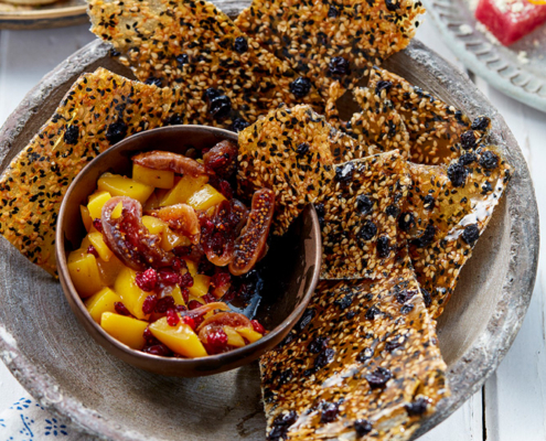 przibylla-fotograf-levante-sesam-gewuerzkrokant-mit-mangokompott Eine Schale mit knusprigen Crackern mit Körnern wird mit einer kleinen Schale mit Mango-Kompott und getrockneten Feigen serviert. Daneben sind ein Teller mit Atayef und eine Schale mit rosafarbenem Konfekt, garniert mit Orangenschale, auf dem weißen Holztisch zu sehen, der mit einem blau gemusterten Tuch dekoriert ist – perfekt für Food-Fotografie.
