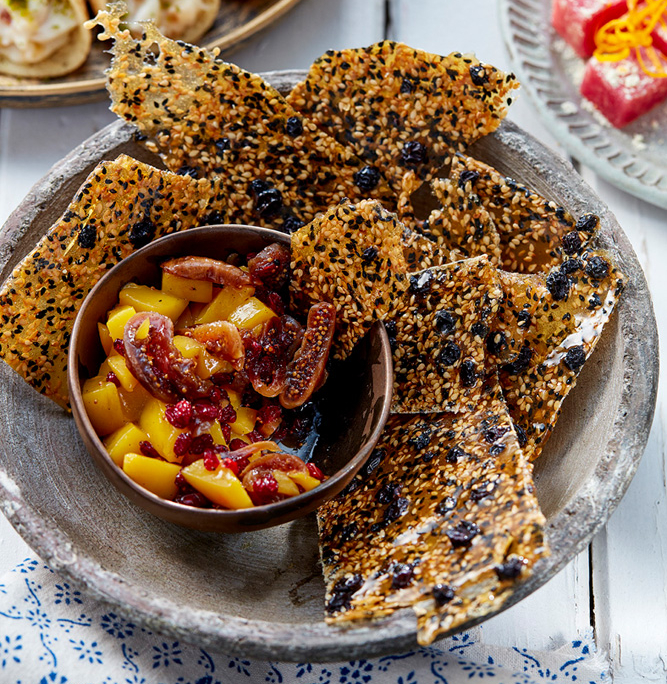 przibylla-fotograf-levante-sesam-gewuerzkrokant-mit-mangokompott Eine Schale mit knusprigen Crackern mit Körnern wird mit einer kleinen Schale mit Mango-Kompott und getrockneten Feigen serviert. Daneben sind ein Teller mit Atayef und eine Schale mit rosafarbenem Konfekt, garniert mit Orangenschale, auf dem weißen Holztisch zu sehen, der mit einem blau gemusterten Tuch dekoriert ist – perfekt für Food-Fotografie.